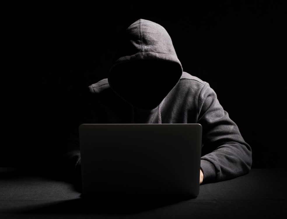 Pessoa encapuzada diante de um notebook em ambiente escuro, representando crimes digitais e o risco de ser vítima de estelionato pela internet.