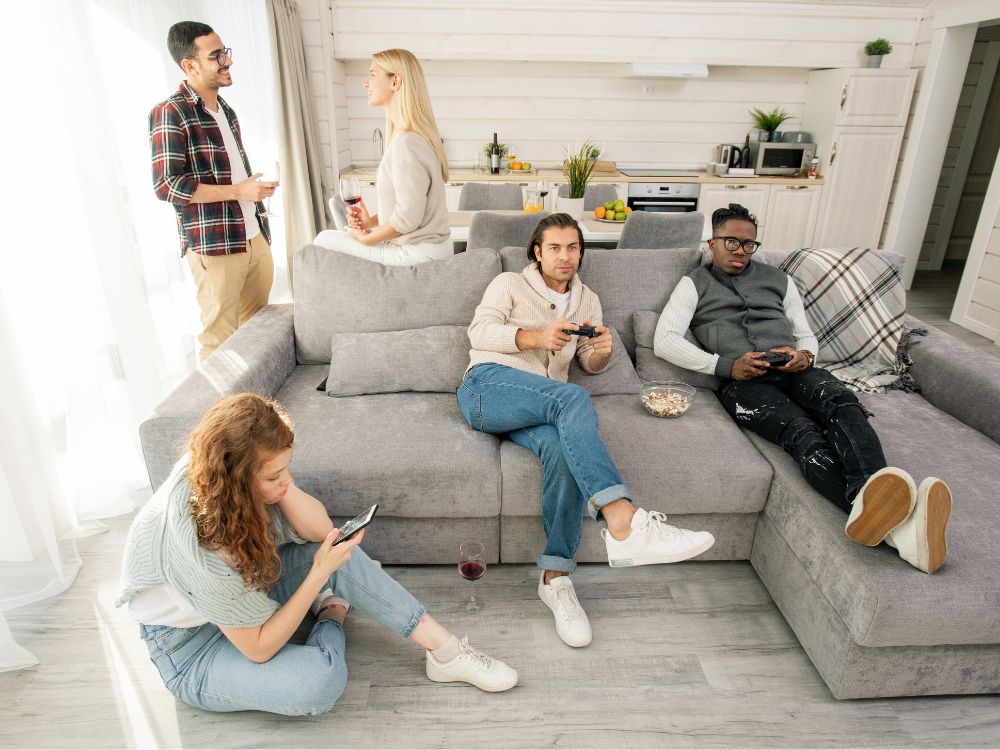 Grupo de jovens em apartamento compartilhado, conversando, usando celular e jogando videogame em ambiente integrado e descontraído.