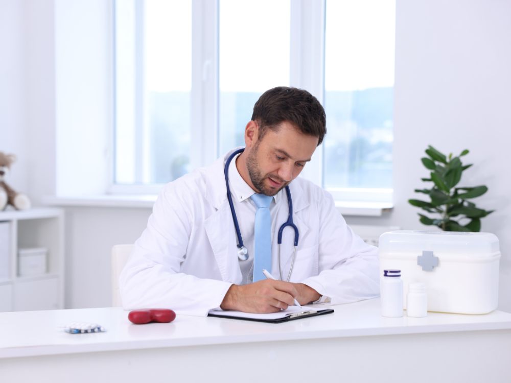 Médico em consultório escrevendo em prontuário durante atendimento, com estetoscópio e materiais clínicos sobre a mesa.