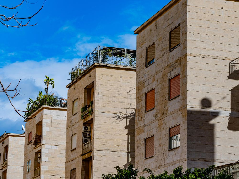 Prédios residenciais de fachada clara com varandas e plantas, sob céu azul em área urbana.